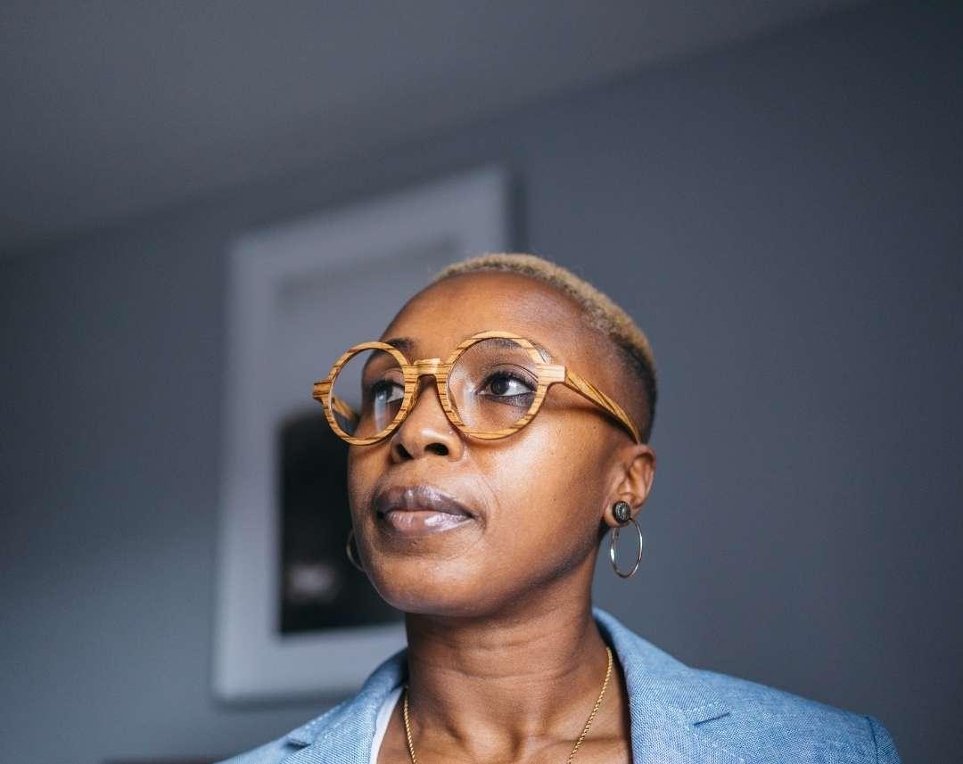 Black woman with short blond hair wearing round wooden glasses made of light brown Zebrawood with subtle black stripes sitting on rosewood wooden case by NURILENS.