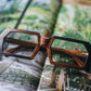 Brown and black rectangular wooden glasses made of black birch and brown kevazingo wood by NURILENS sitting on a tropical magazine page.
