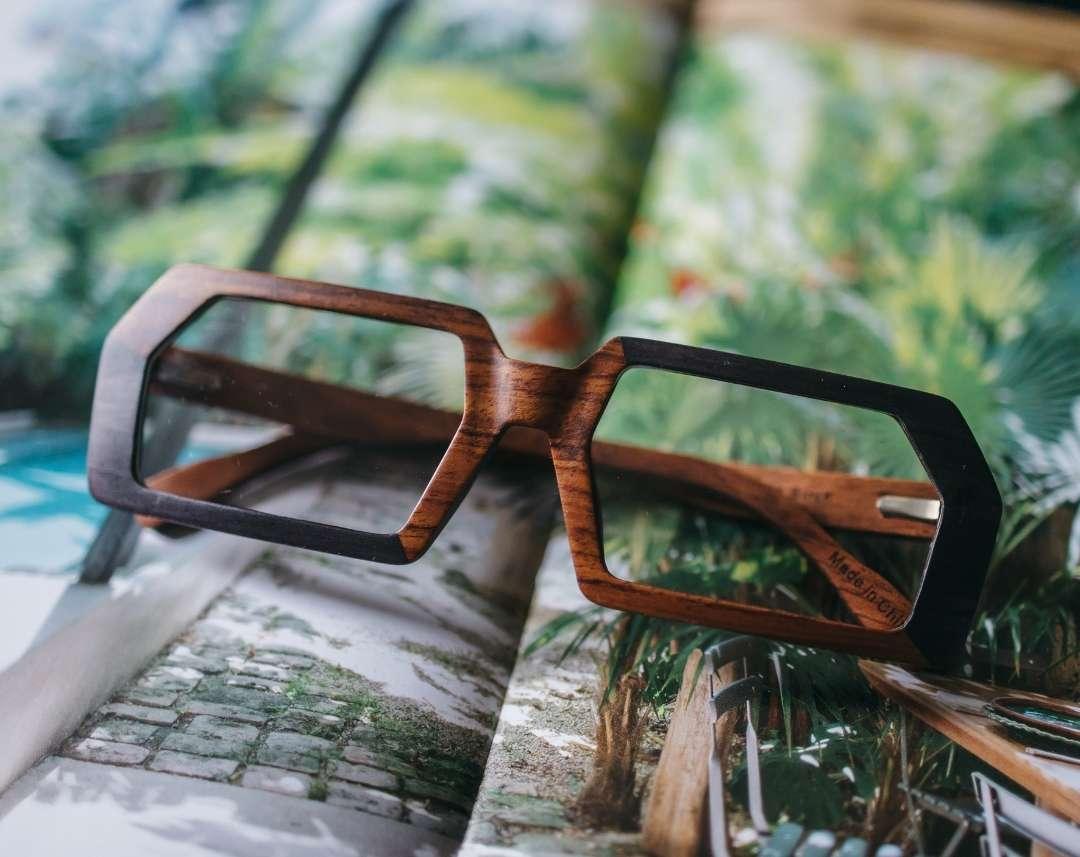 Brown and black rectangular wooden glasses made of black birch and brown kevazingo wood by NURILENS sitting on a tropical magazine page.